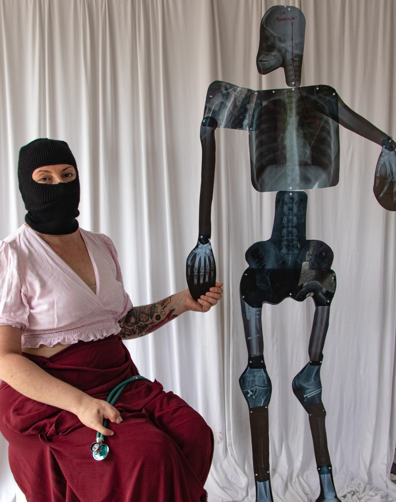 Foto de Nathi, sentada em um banco e Martín, El periodista em pé. Nathi usa um capuz preto onde só aparecem os olhos, blusa rosa claro até os cotovelos e saia longa vermelha, no seu colo um estetoscópio verde. Martín, el periodista é uma criatura humanóide, mostra os ossos com folhas de Raio X. Na cabeça a frase “La Revolução”, uma linha vai descendo pelo pescoço e chega até a parte dos pulmões e outra frase aparece “Resiste Corazón”. Estão de mais conectadas. O fundo é todo branco.